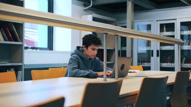 Concentrated student typing on a laptop computer while studying at a desk in a public library.