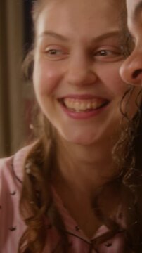 Vertical shot of Caucasian teenager girl smiling while friend applies makeup and cheek blusher with brush during slumber party. Cosmetic beauty product commercials and teenage lifestyle marketing