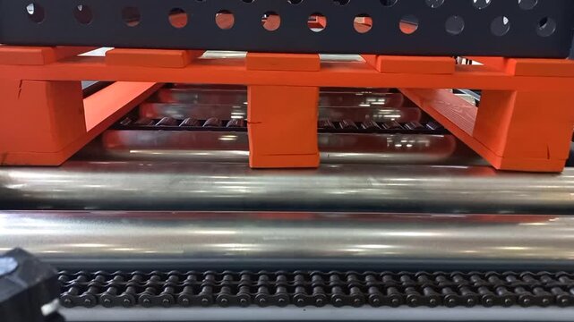 An orange wooden pallet moves along an automated industrial roller conveyor with metal cylinders and drive chains inside a warehouse facility, captured in a close-up view during an indoor process.