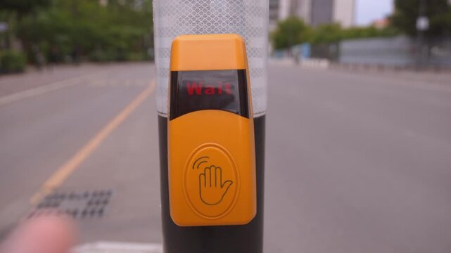 Closeup of a person's hand pressing a yellow pedestrian call button at a crosswalk to activate the traffic signal, with cars passing in the background on a city street