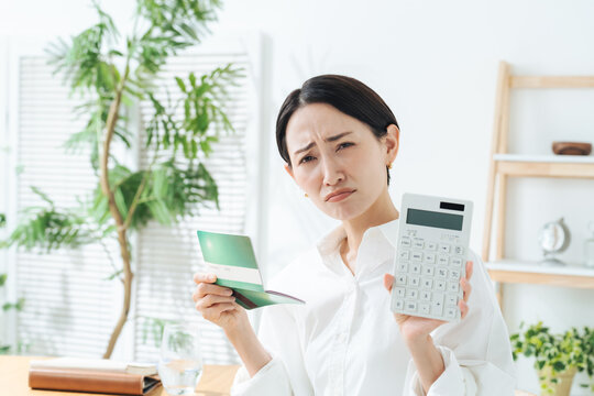 通帳と電卓を持って悩む女性。貯金、マネー、投資、資産運用、不満、疑問、悩み、不安、心配、ローン、借金