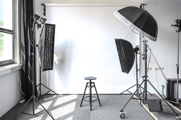 Small photo studio with a natural light from a window, white paper backdrop, lamps on tripods with softbox, striplight and umbrella, and an adjustable black stool, copy space © Maren Winter