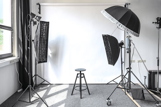 Small photo studio with a natural light from a window, white paper backdrop, lamps on tripods with softbox, striplight and umbrella, and an adjustable black stool, copy space