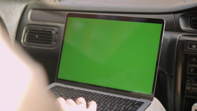 Person uses laptop with green screen inside car. Hands type on keyboard, focused on screen. View from behind, showing car dashboard and controls. Ideal for remote work, travel, or tech-themed content