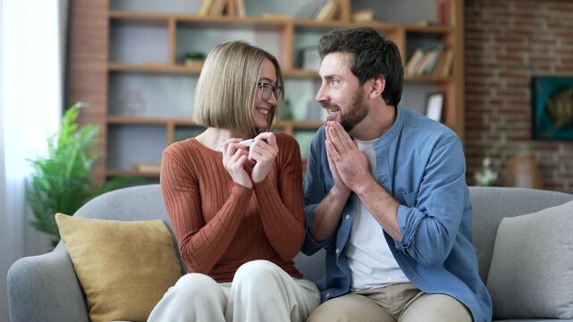 Smiling married couple are happy with a positive pregnancy test hold in hands sitting on couch in living room at home. Joyful excited family husband and wife celebrate successful outcome and pregnancy
