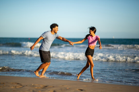 Playful young couple holding hands and running along sunny beach