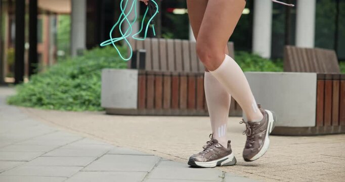A young beautiful female athlete in sportswear with a skipping rope goes for a workout in an urban park among high-rise buildings - legs close-up