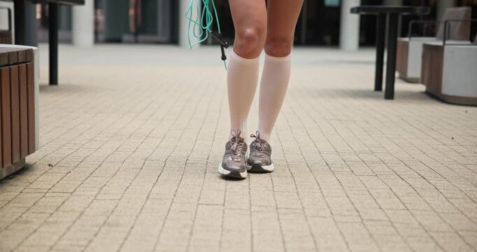 A young beautiful female athlete in sportswear with a skipping rope goes for a workout in an urban park among high-rise buildings - legs close-up