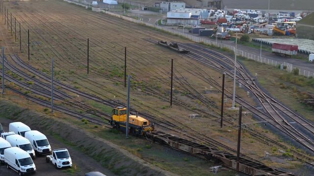 Yellow railway locomotive standing stationary at an industrial railway yard, with hazard warning lights flashing, situated near a cargo storage area.