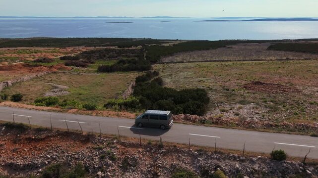 Camper van driving past bushes and rocks with the Adriatic sea in the background. Aerial car follow