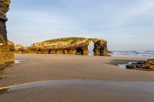 A horizontal scenic view of a monumental natural rock arch and cliffs on the sandy shore of Playa de las Catedrales in Ribadeo, Spain, during low tide at golden hour
