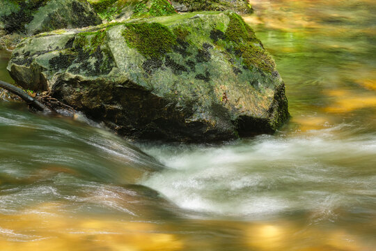 A large boulder in a fast-flowing river in a forest in southern France on a sunny summer day. Image with motion blur in the water and copy space. Metaphor for steadfastness in emotions.	