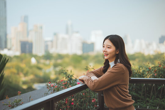 Side view of happy Asian woman smiling and looking away while leaning on railing at rooftop garden, beautiful young female traveler relaxing with city skyscraper background for urban lifestyle, real e