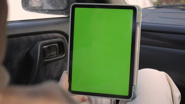 Person holds tablet with green screen inside car. Focus on device, hands, car interior. Calm, neutral expression while viewing screen. Ideal for advertising, app demos, or instructional videos