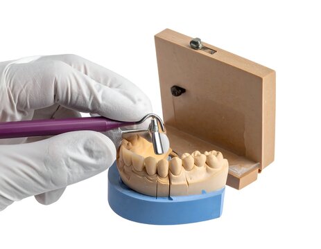 A gloved hand holds a dental tool near a model of upper teeth on a blue base with a wooden case open in the background