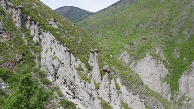 Hoodoo rock pillars with mushroom capstones rising from eroded cliff in lush green mountain valley beneath overcast sky, showing rugged layered stone formations and steep slopes