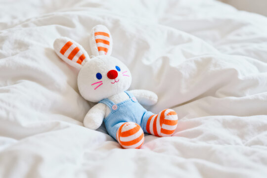 A plush rabbit toy with striped ears and overalls rests on wrinkled white bed linens in a bright indoor space.