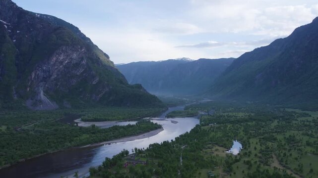Aerial river valley winding through steep forested mountains beneath overcast sky, with broad S-shaped waterway, distant snowcapped peaks, rugged cliffs, and vast untouched wilderness