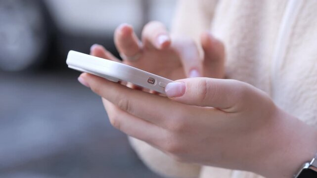 Close-up of hands holding white smartphone. Fingers tap screen with focus. Background blurred, showing casual setting. Person wears light sweater and watch. Ideal for tech, communication