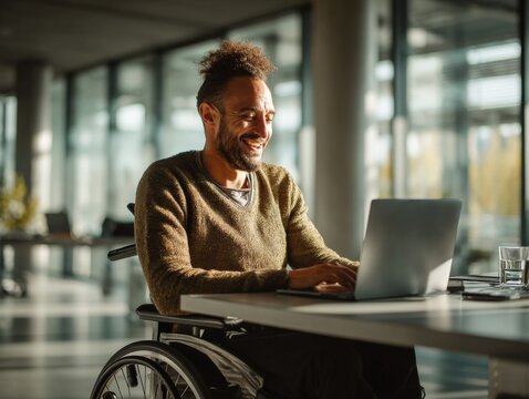 Smiling man in wheelchair working on laptop in modern office with natural light