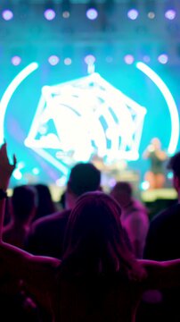 vertical video. A woman dances with her arms raised in front of a large stage at an awesome rock festival. A person dances in the crowd with an arm raised at a live concert. Blurred video.