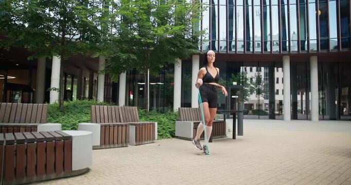 Young beautiful woman athlete in sportswear training and jumping rope in an urban park among high-rise buildings and trees