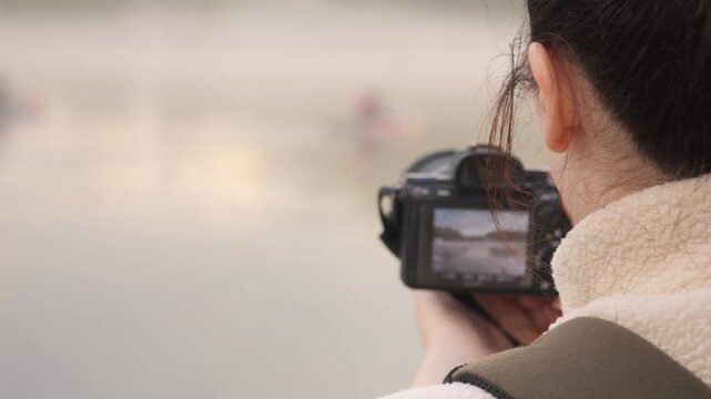 Person holds camera, focusing on distant water scene. Focus on back of head, showing intent to capture. Soft, blurred background suggests peaceful outdoor setting. Could use for travel, nature