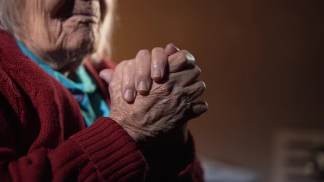 In a dimly lit room, a view of an elderly woman fingers interlaced in prayer