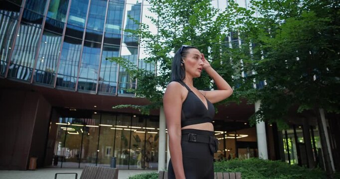 Young beautiful female athlete in sportswear trains in an urban park among high-rise buildings and trees and rests during a break between exercises