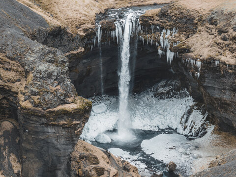 Aerial view of a waterfall cascading into a rocky gorge surrounded by ice and snow in the rugged landscape of Reykjavik, Reykjavikurborg, Iceland.