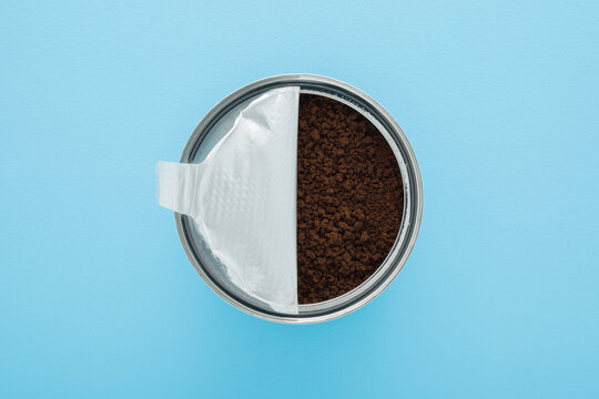 Opened can with dry dark black granules of instant coffee on light blue table background. Pastel color. Closeup. Flat lay. Top down view.