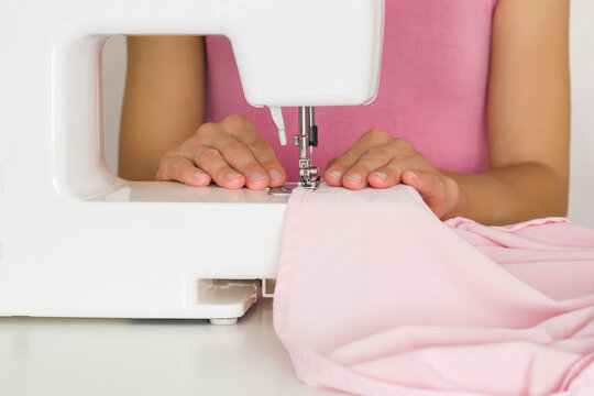 Young adult woman hands working with white sewing machine and sewing female dress from light pink gabardine elastane fabric. Closeup. Front view.