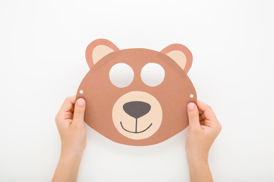 Little child boy hands holding brown bear face mask from application paper on light gray table background. Closeup. Point of view shot. Flat lay. Top down view.