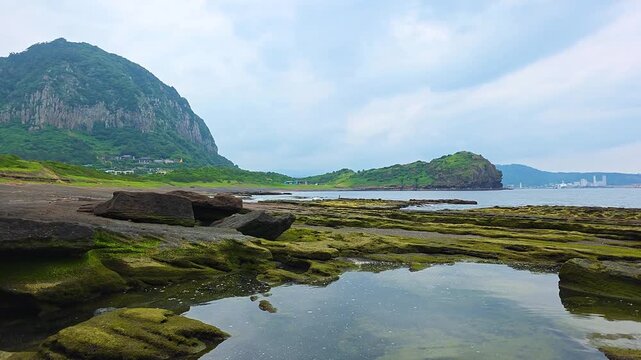 Sanbangsan Mountain and Yongmeori Cliff rise above green algae covered Hamori Layer tuff platform with calm tide pools at low tide on Sagye Beach Jeju Island South Korea