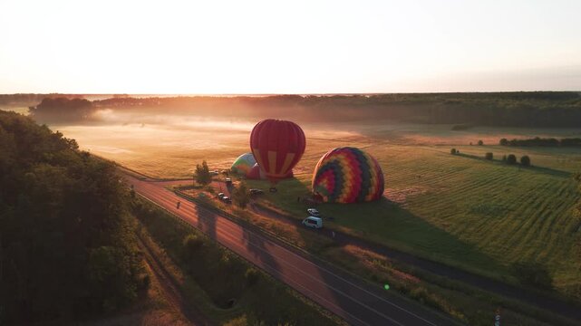 Cinematic aerial shot of colorful hot air balloons inflating in a rural field during a misty sunrise. Great for travel, adventure, and wellness content.