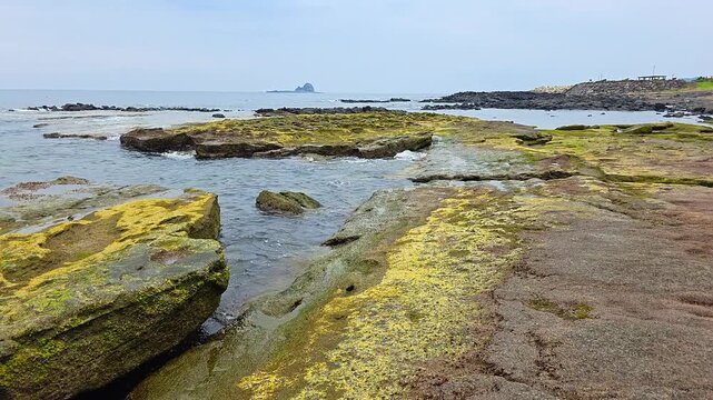 Moss-covered tuff and sandstone formations along Sagye Beach, with ocean waves washing over layered volcanic rocks on Jeju Island