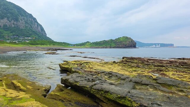 Layered volcanic tuff and sandstone rocks along Sagye Beach bay, with Sanbangsan Mountain and small islet in background on Jeju Island