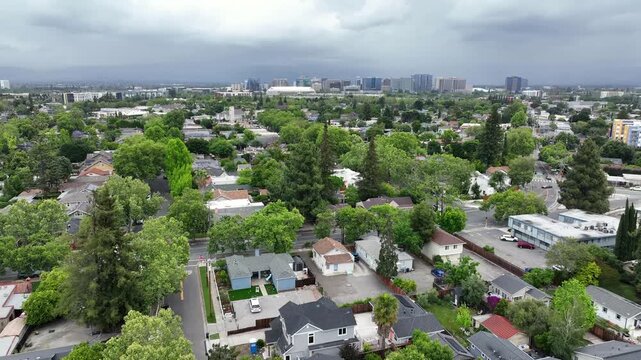 Dense tree-lined residential neighborhood with mixed rooftops stretches toward downtown San Jose skyline beneath dramatic overcast storm clouds rolling. California, USA. Aerial drone wide shot