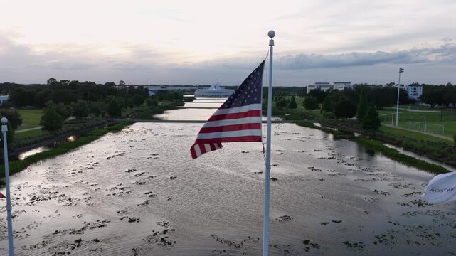 American and Florida state flags wave above tranquil canal, futuristic domed campus building rising beyond tree-lined waterway at golden dusk. Lakeland, Florida, USA. Aerial drone orbit shot.