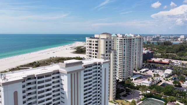 High-rise beachfront condominium towers overlook sandy shoreline and turquoise gulf waters, tennis courts and pool nestled below palm trees. Clearwater, Florida, USA. Aerial drone flyover shot