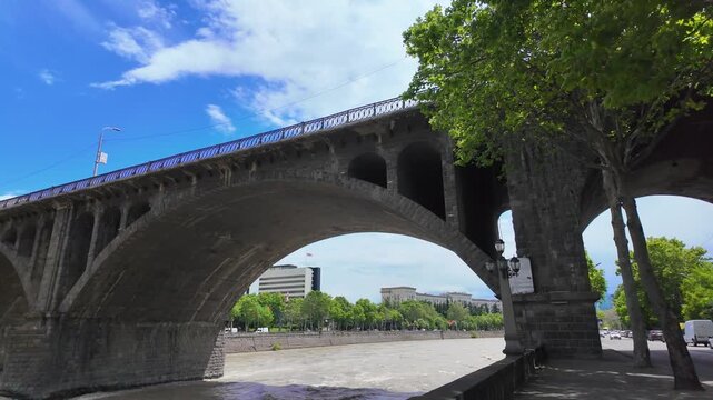 Personal perspective view walking along the embankment under the historic galaktioni bridge, which spans the muddy mtkvari river in the beautiful city of tbilisi, georgia, on a sunny day