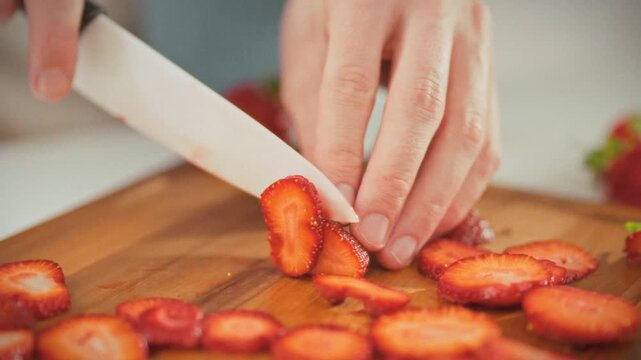 In a bright kitchen, a man slices ripe red strawberries into thin slices on a cutting board, preparing the juicy berries for serving. Concept of making a homemade dessert with fresh summer berries
