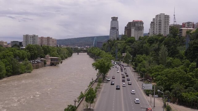 Aerial view of cars and vans driving on a busy highway along the embankment of the muddy mtkvari river in tbilisi, the capital of georgia, surrounded by lush green trees and city buildings