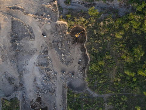 Aerial view of a sand and stone quarry featuring an excavator loading trucks on a dirt path, surrounded by rocky terrain and dense vegetation in Manisrenggo, Central Java, Indonesia.
