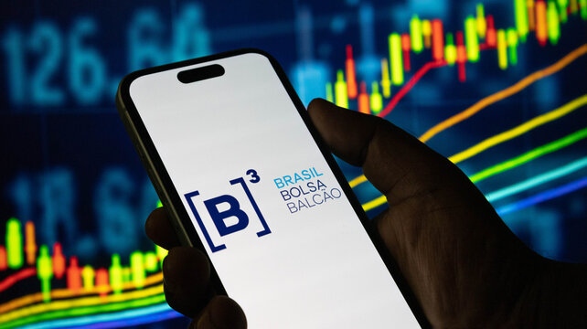 Hand holding smartphone with b3 brasil bolsa balcao logo over digital financial indicators.