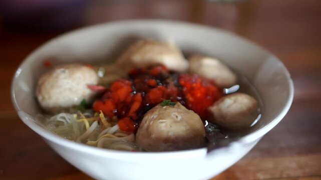 A bowl of traditional Indonesian beef meatballs with noodles, rice vermicelli, savory broth, chili sauce, and sweet soy sauce.