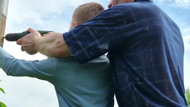 Father guiding his young son to use cordless drill outdoors while preparing materials for future fence, hands-on learning experience, father-son bonding, practical skills training.