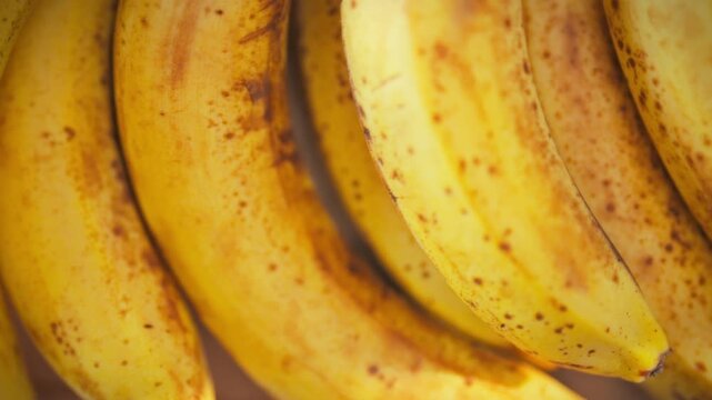 A close-up of a bunch of yellow bananas with dark spots on the peel, indicating ripeness. The camera moves smoothly from above, sunlight falling on the surface. Ripe summer fruit concept