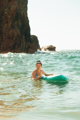 Obraz premium beach Ursa, Portugal. woman is in water holding surfboard while smiling. The ocean is bright and reflecting sunlight. Rocks are visible in the background near the shore.