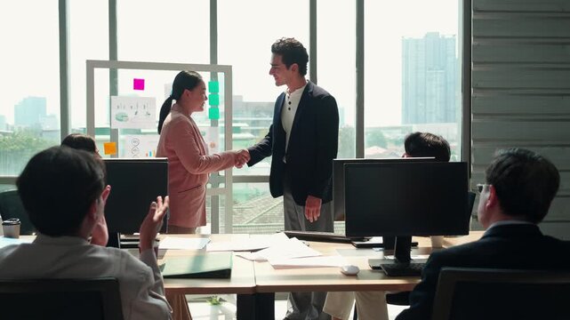 Professional businessman shaking hands with asian businesswoman during office meeting, man and partnership colleagues sitting at computer desk clapping hands with celebrating success business deal.